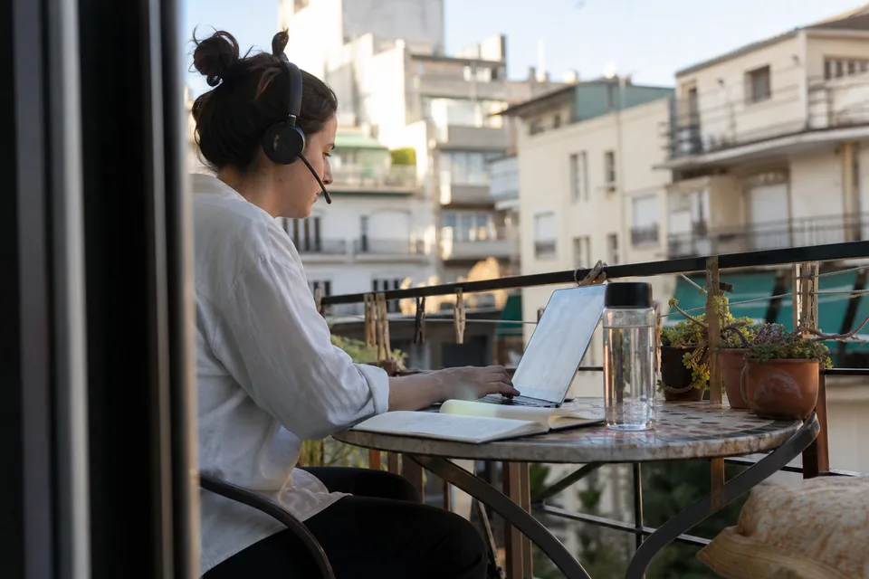 Adult woman with a headset working on a laptop at a small balcony table in the morning, notebook and plants beside her