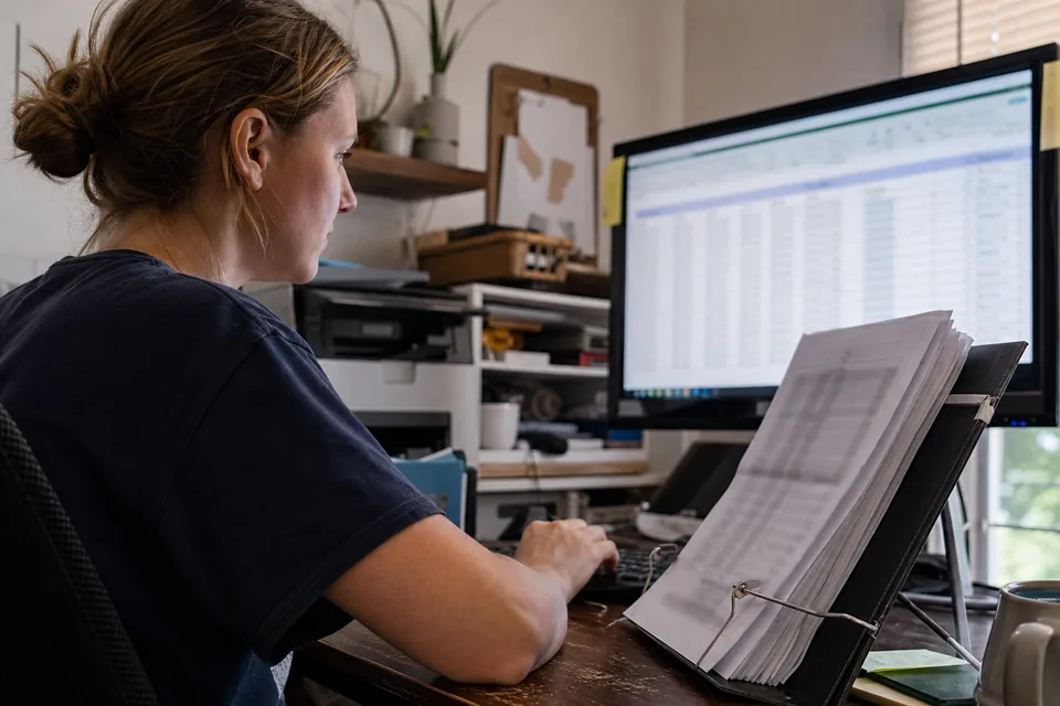 Data entry assistant typing figures from paper forms held on a document stand into a spreadsheet