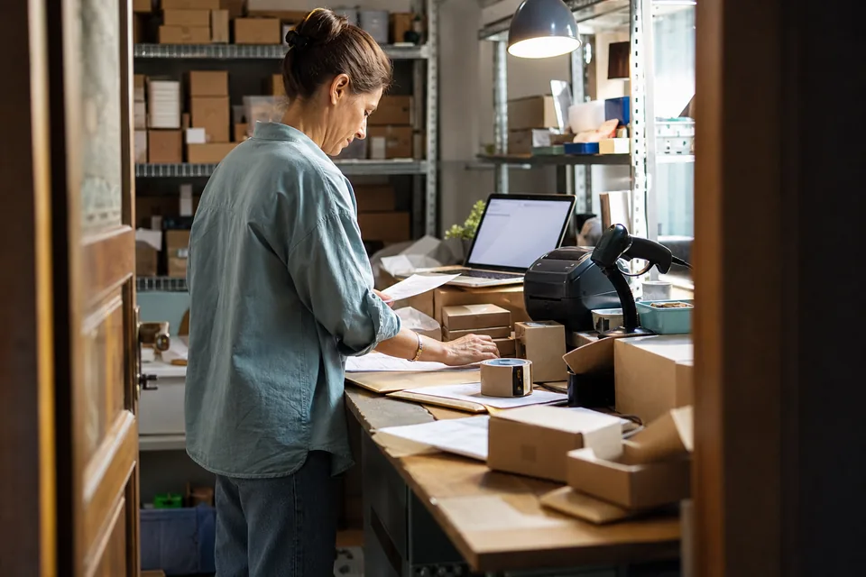 Adult woman assistant at a packing table in an ecommerce back room with order printouts, label printer, boxes and a laptop showing an order list