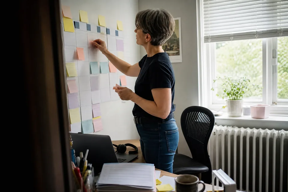 Adult woman assistant at a home desk moving sticky notes on a weekly wall planner, a laptop and a headset on the desk