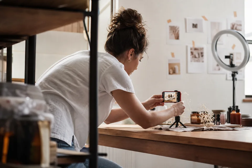 Social media assistant photographing a product flat lay with a phone on a small tripod beside a printed content calendar