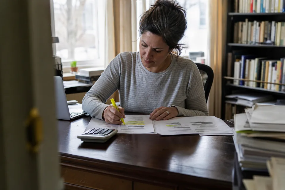 Adult woman comparing two printed quotes with a highlighter and a calculator at a home office desk