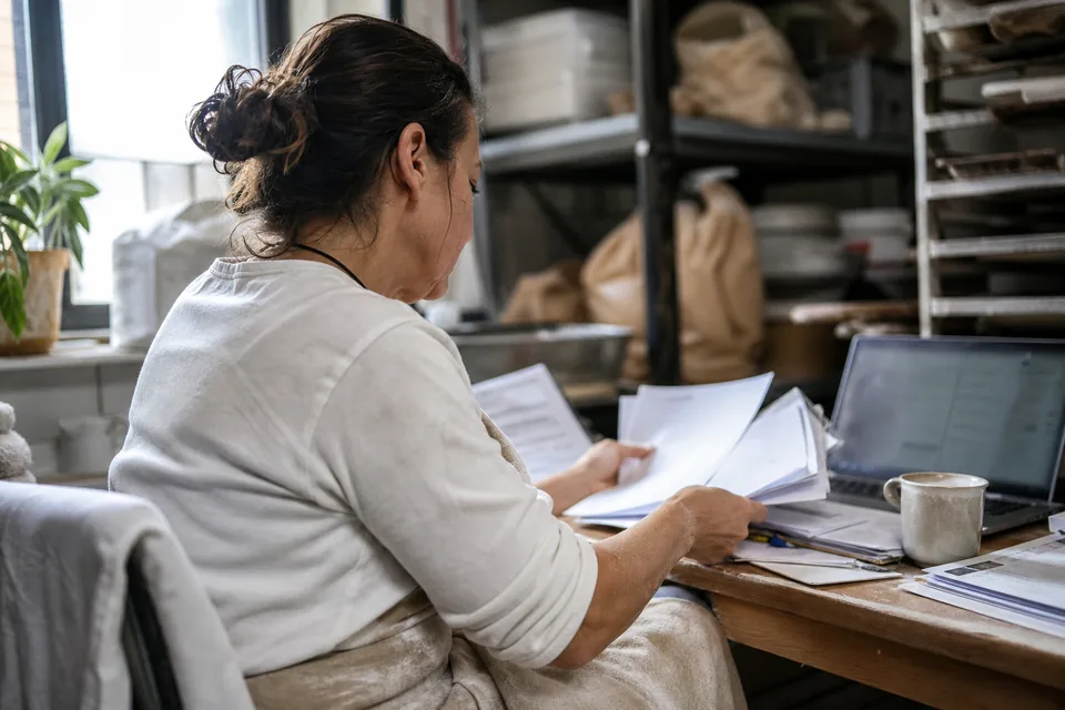 Bakery owner in an apron sorting invoices in a cramped back office with a laptop open beside her