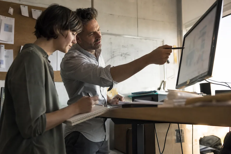Founder at a standing desk in a small office pointing at a monitor while a colleague takes notes, printed pages pinned on a corkboard behind